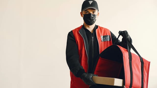 Delivery man wearing a mask and gloves with a red thermal bag and box indoors.