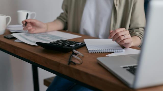 A woman manages finances at home, using a laptop and calculator on a wooden desk.