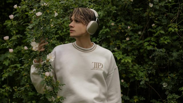 A young man wearing headphones and a white sweatshirt stands amidst lush greenery and flowers.