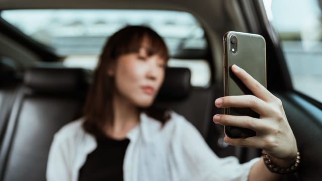 Confident Asian female in casual clothes sitting in car passenger seat and taking selfie on modern mobile phone