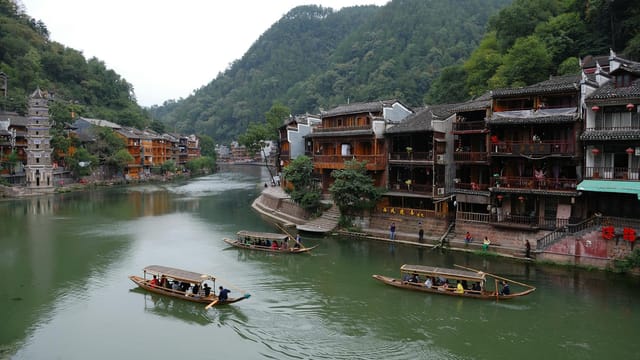 Traditional wooden boats on a serene river beside historic Chinese architecture and lush mountains.