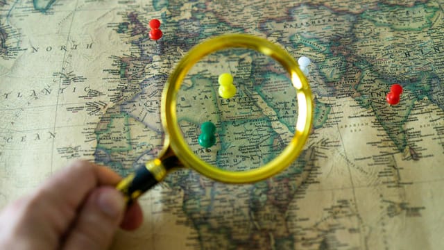 Close-up of a hand holding a magnifying glass over a world map with colorful pins indicating travel destinations.