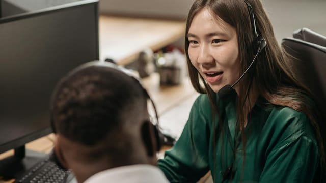 Young Asian woman in a call center talking with a colleague in an office environment.