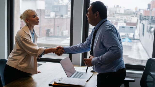 Young female candidate shaking hands with employer before job interview in light office with urban landscape through windows