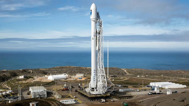 A SpaceX rocket stands on a launch pad overlooking the ocean under a clear sky.