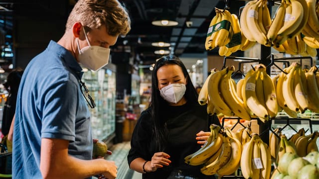 Masked shoppers examining bananas in an urban supermarket during the COVID-19 pandemic.