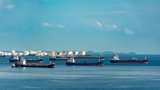 A fleet of cargo ships docked near oil storage tanks along a serene coastline with a clear blue sky above.