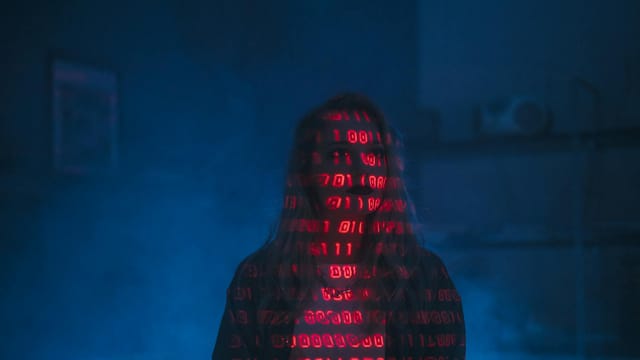 A mysterious silhouette with red binary code projected over the face, set against a dark, moody background.