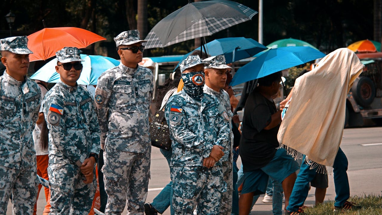 Military personnel stand with umbrellas in a street, showing resilience during a rainy event.