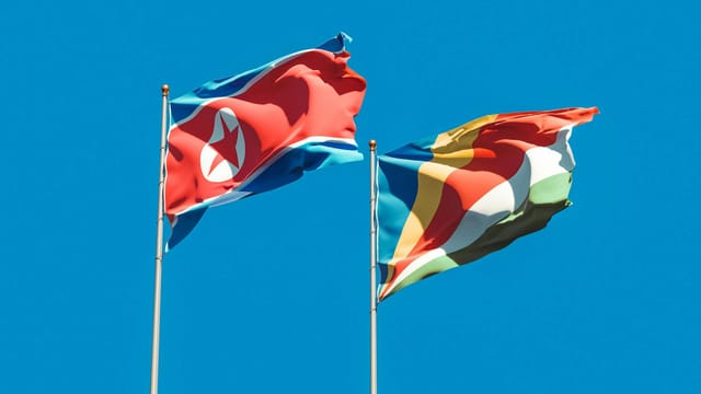 Vibrant national flags of North Korea and Seychelles on flagpoles waving under a clear blue sky.