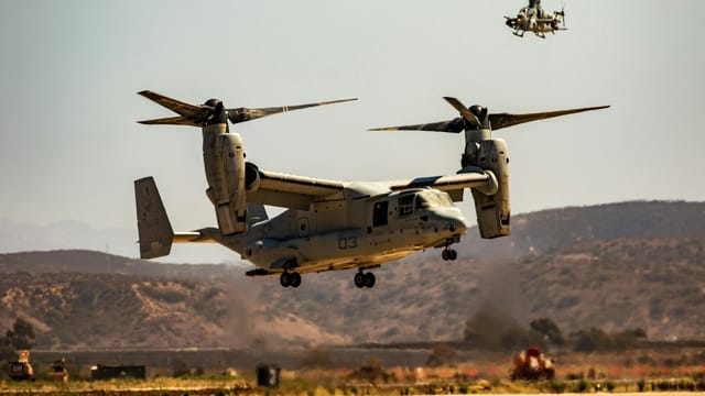 A military Osprey aircraft alongside a helicopter flying over arid landscape in daylight.
