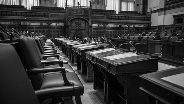 Black and white photo of a traditional legislative assembly chamber, featuring empty seats and desks.
