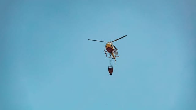 Helicopter carrying water bucket for firefighting against a clear blue sky.