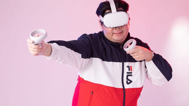 Adult man wearing VR goggles and jacket, playing with controllers against a pink studio background.