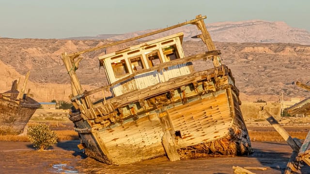 Derelict wooden boat in a dry coastal area in Bushehr, Iran, under warm sunlight.
