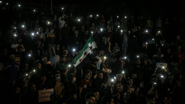 A large group of people gather at night in Syria, holding flashlights and the Syrian flag, symbolizing unity.