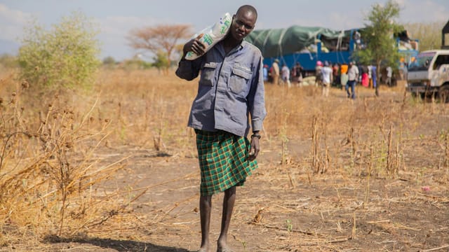 A man carries a bag in a rural African landscape, with a truck and people in the background.