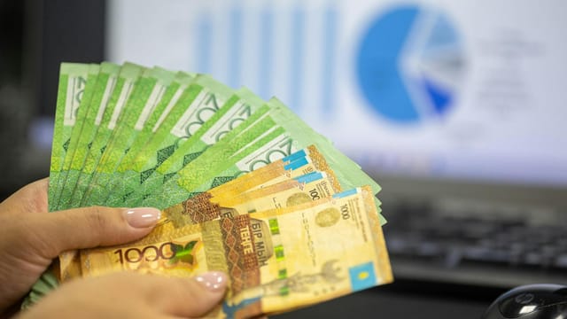 Close-up of hands holding Kazakhstani tenge bills with financial charts in background.