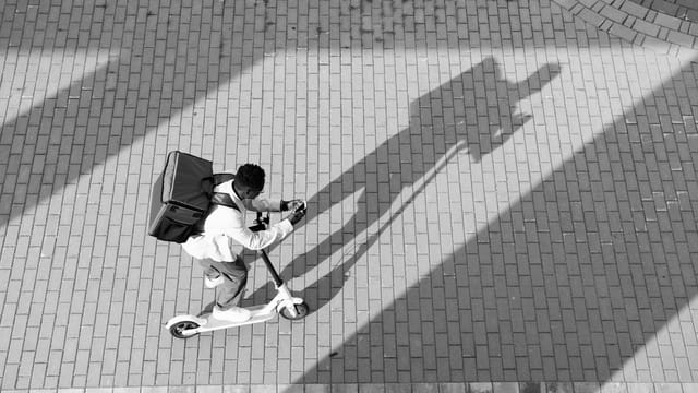 A courier on a scooter travels over a sunlit brick path in a monochrome urban scene.