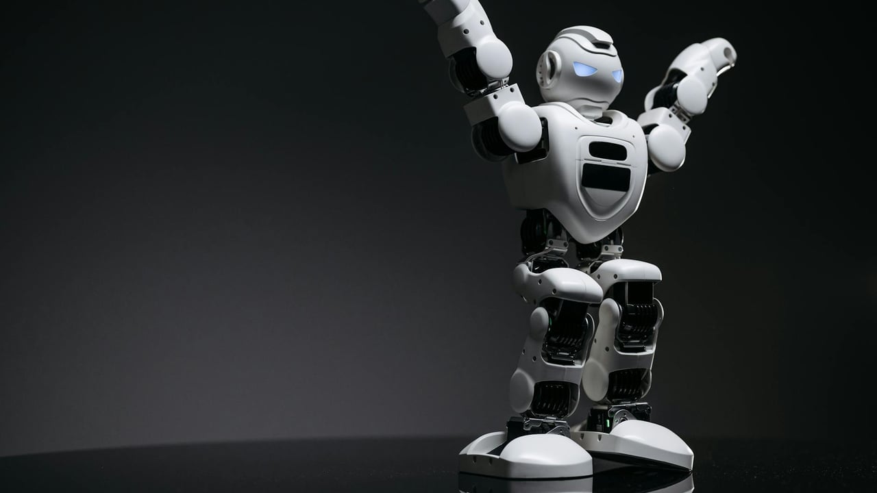 Close-up studio shot of a white robot toy with LED eyes raised in victory on a gray background.