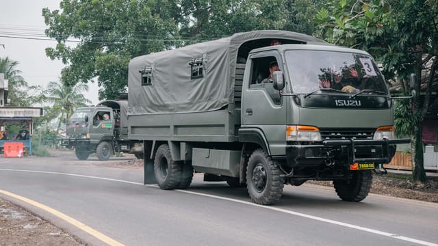 Military trucks traveling on a rural road surrounded by trees and local buildings.