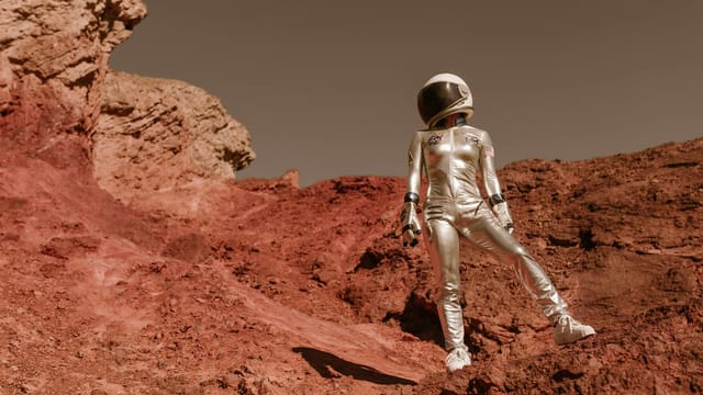 Astronaut in space suit standing on red rocky terrain resembling Mars' surface.