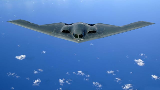 A stealth bomber aircraft flying over a clear blue ocean with scattered clouds.