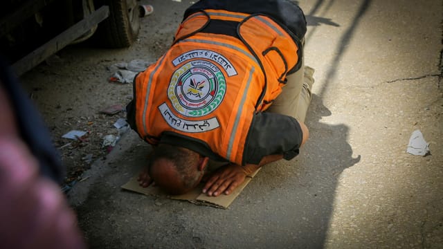An emergency worker in an orange vest prays on a street in Gaza, depicting a moment of faith amidst turmoil.