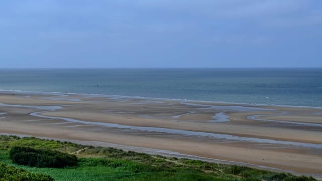 Tranquil view of Omaha Beach in France, showcasing vast sand and calm sea under blue skies.