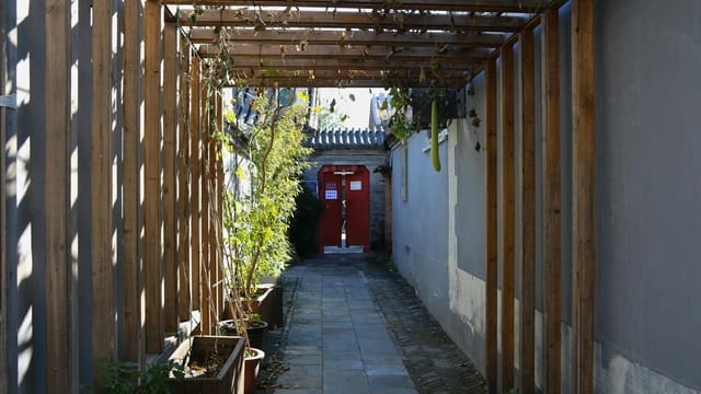 A narrow alley in Beijing's hutongs with red doors and wooden trellis casting shadows, symbolizing traditional Chinese architecture.