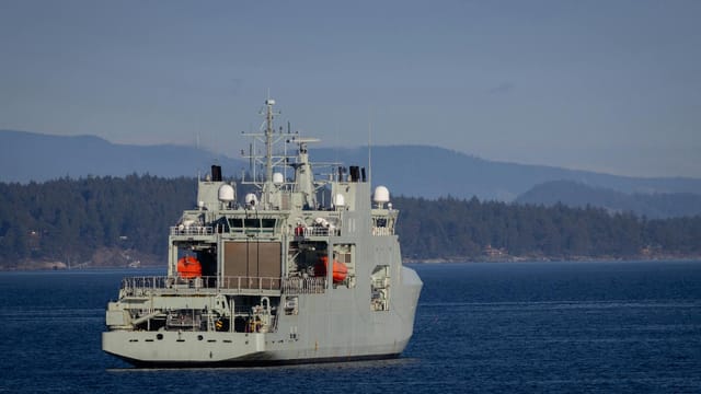 A military warship navigates through open waters with a coastline backdrop on a clear day.