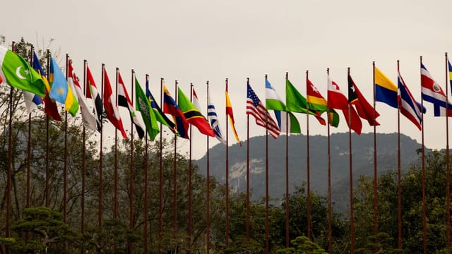 A display of various national flags on flagpoles set against a mountain backdrop, symbolizing global unity.