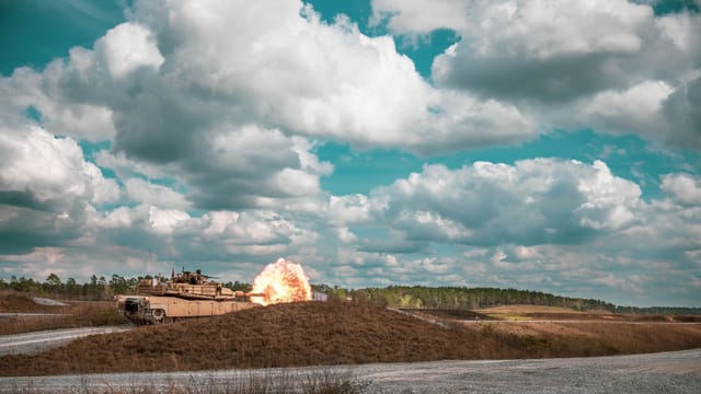 A military tank fires a shot in an open landscape under a dramatic sky.