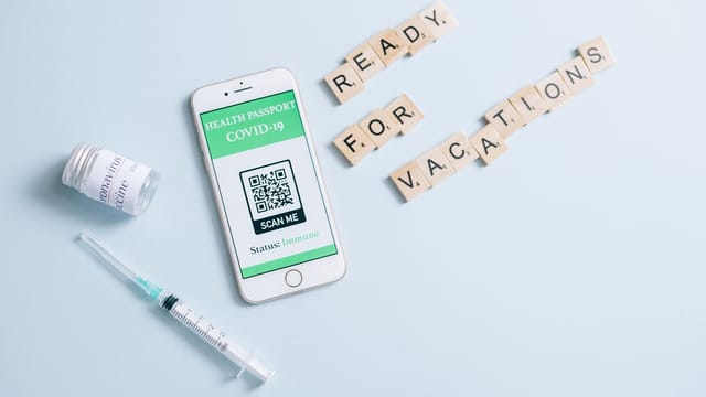 Smartphone displaying COVID-19 health passport with scrabble tiles spelling 'Ready for Vacations', vaccine vial, and syringe.