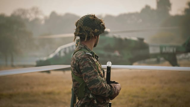 An Indian soldier stands guard in a field with a military helicopter in the background.