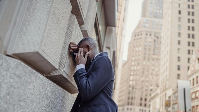 A man in a suit leans against a city wall, seemingly overwhelmed.