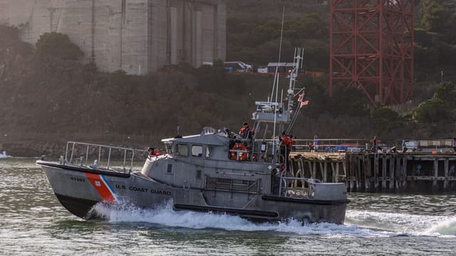 US Coast Guard boat navigating near a pier with coastal scenery in the background.