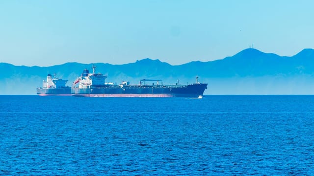 A large container ship sails offshore with mountains in the background under a clear blue sky.