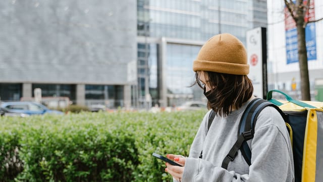 Woman with thermal bag checking phone in urban setting, delivering food.