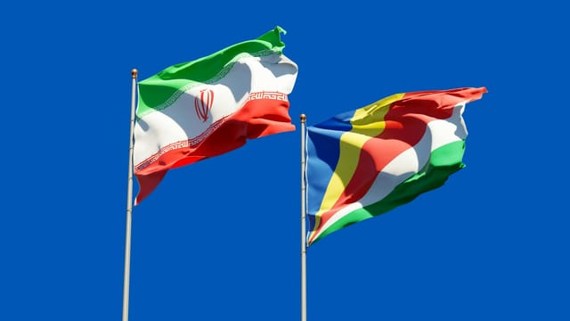 National flags of Iran and Seychelles waving on poles against a bright blue sky, symbolizing international relations.