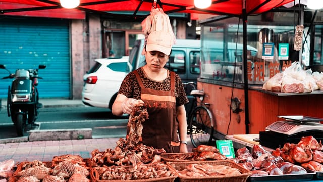 A vendor at a street market selling various meats under a red tent with urban background.