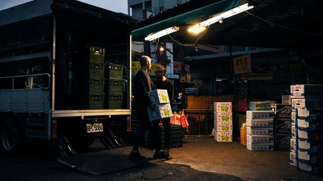 Workers unloading boxes from a delivery truck at an urban market during dusk.