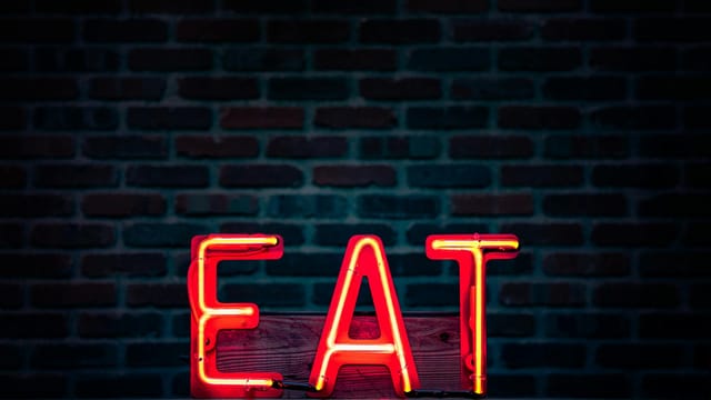 Vibrant neon 'EAT' sign in red glowing boldly against a dark brick wall background.