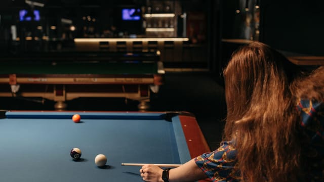A woman with red hair playing billiards in a dimly lit pool hall, aiming with a cue stick.