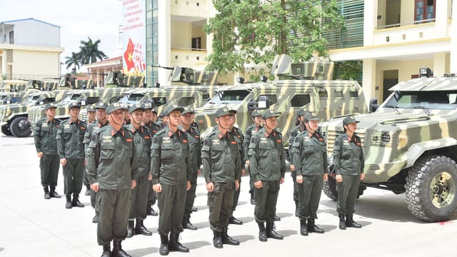 Group of military personnel in uniform stands in formation near armored vehicles on a sunny day.
