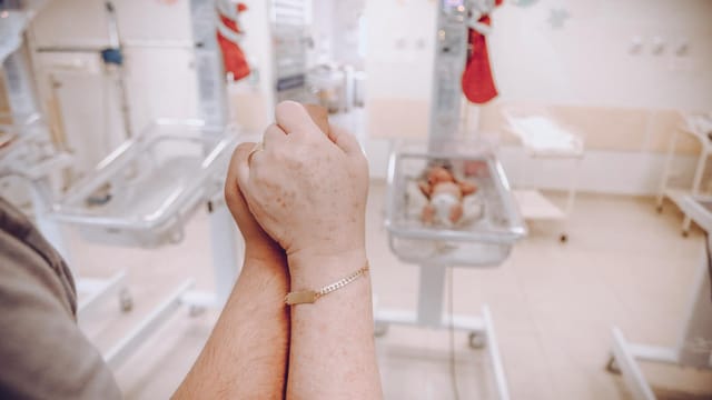 A parent's hands clasped together in a hospital nursery, with newborns in the background.