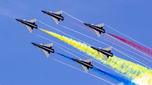 Six jet fighters perform an aerial display with colorful smoke trails against a clear blue sky.