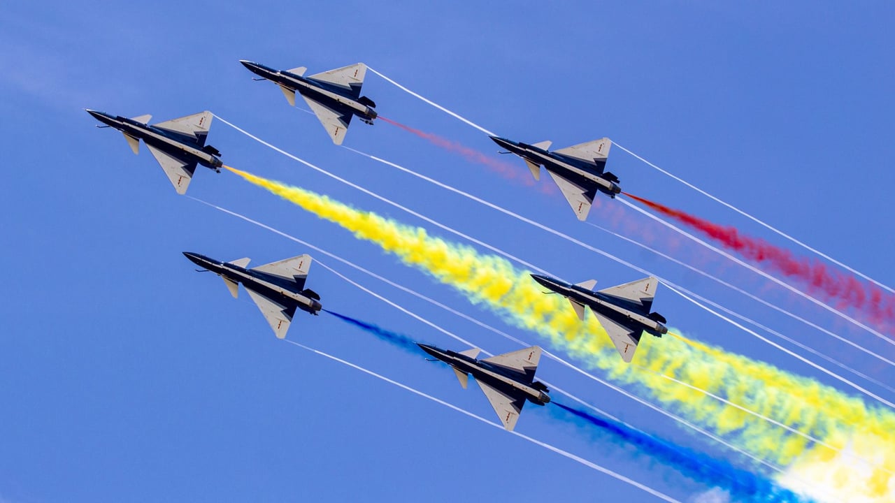 Six jet fighters perform an aerial display with colorful smoke trails against a clear blue sky.