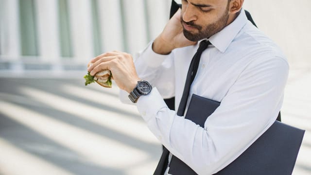 Professional man in corporate attire checking time while holding a sandwich outdoors.