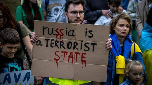 A group of adults and children holding a protest sign in an outdoor rally against Russia's actions.